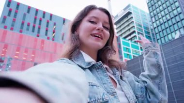 Kot ceketli şık genç bir kadın modern şehir binalarının arka planına karşı özgüvenli bir pozla selfie çekiyor. Canlı ve çağdaş bir şehir yaşam tarzı anı.