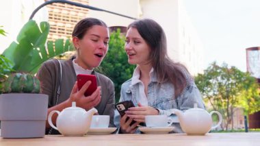 İki kadın dışarıda bir masada oturuyor, kahvenin tadını çıkarıyor ve cep telefonlarını kullanıyorlar. Modern ve rahat bir atmosferde, açık hava yeşiliyle birlikte gülümsüyor ve anları paylaşıyorlar..