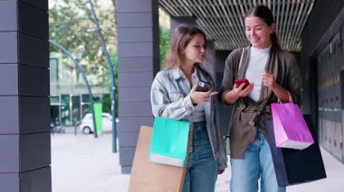 Alışveriş torbaları taşıyan ve dışarıda yürürken akıllı telefon kullanan iki genç kadın. Şehir ortamında arkadaşlığın, stilin ve modern alışveriş deneyimlerinin sergilendiği gündelik ve neşeli bir an.. 