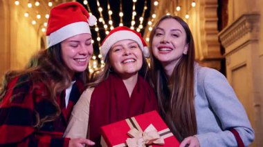 Noel 'i dışarıda Noel Baba şapkası takarak kutlayan üç mutlu arkadaş. Bir hediyeyi kurdeleyle tutarken sevinçle gülümserler. Arka planda, bayram ruhunu yakalayan parlak şenlik ışıkları vardır.