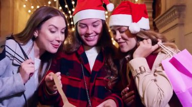 Noel Baba şapkası takan üç neşeli kadın, tatil sezonunda güzel Noel ışıklarının önünde gülümserek selfie çekiyorlar. Birlikte keyifli bir an geçiriyorlar. Şenlikli şehirde kutlama yapıyorlar..