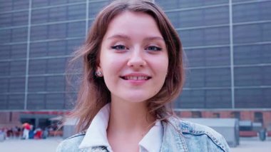 Kot ceketli şık genç bir kadın modern şehir binalarının arka planına karşı özgüvenli bir pozla selfie çekiyor. Canlı ve çağdaş bir şehir yaşam tarzı anı.