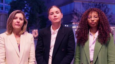 Üç profesyonel kadın, gece bir şehir ortamında kollarını kavuşturarak güvenle ayakta duruyor. İş kıyafetleri içinde, güç, kararlılık ve takım çalışması yayarlar..
