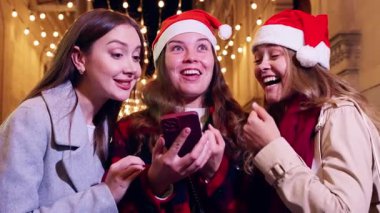 Üç kadın Noel Baba şapkası takıyor, gülümsüyor ve telefonla gülüyor, ellerinde alışveriş torbalarıyla bir festival caddesinde. Sahne, tatil sezonunun neşe ve heyecanını yakalıyor..