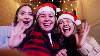 Noel Baba şapkası takan üç neşeli kadın gülüyor ve Noel gecesini kutluyor, ellerinde hediyeler ve şehrin şenlikli ışıklarının önünde poz veriyorlar. Neşeli, coşkulu bir arkadaşlık ve tatil ruhu anı..