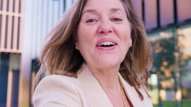 Bej ceketli neşeli bir kadın, saçları rüzgarda uçuşan parlak bir şekilde gülümsüyor. Kentsel bir arkaplanda yakalandığında, kendine güven, pozitiflik ve mutluluk yayar.. 