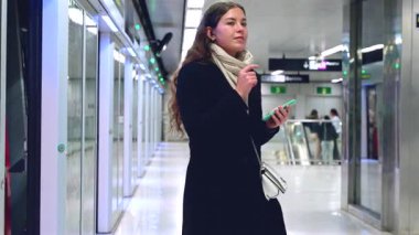  Palto ve eşarp giyen şık genç bir kadın akıllı telefonu ve kablosuz kulaklıklarını kullanarak metro istasyonunda yürüyor. Kendine güveni tam ve şehir ortamında yaşıyor.. 