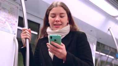 Siyah paltolu ve bej eşarplı genç bir kadın modern bir metro treninin içinde elinde akıllı bir telefonla metal bir direği kavrıyor. İşe gidip gelirken odaklanmış görünüyor. Elinde şık beyaz bir çanta var.. 