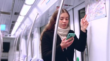  Siyah ceketli ve bej eşarplı odaklanmış genç bir kadın akıllı telefonunu tutarken metro haritasını kontrol ediyor. Modern bir metro treninde rotasını çiziyor..