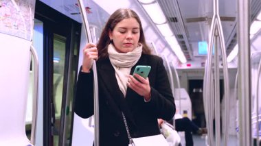 Siyah paltolu ve bej eşarplı genç bir kadın modern bir metro treninin içinde elinde akıllı bir telefonla metal bir direği kavrıyor. İşe gidip gelirken odaklanmış görünüyor. Elinde şık beyaz bir çanta var.. 
