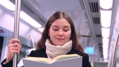 Modern bir metro treninde duran siyah paltolu ve bej eşarplı odaklanmış genç bir kadın metal bir direğe tutunarak kitap okuyor. Seyahat sırasında sakin bir anın tadını çıkarıyor..