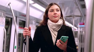 Siyah paltolu ve bej eşarplı genç bir kadın modern bir metro treninin içinde elinde akıllı bir telefonla metal bir direği kavrıyor. İşe gidip gelirken odaklanmış görünüyor. Elinde şık beyaz bir çanta var.. 