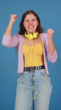 Boynuna sarı kulaklıklar takmış, sevinçle kutlama yapan, yumruklarını kaldıran, gülümseyen ve mutluluğunu ifade eden bir kadın stüdyo ortamında mavi bir arka planda sıradan kıyafetler giyiyordu.