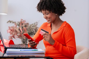 Afrikalı Amerikalı bir öğrenci evde telefon kullanırken kredi kartıyla online ödeme yapıyor. Kitaplı, kırtasiyeli ve şık dekorlu modern çalışma alanı. E-ticaret, dijital işlem ve finans