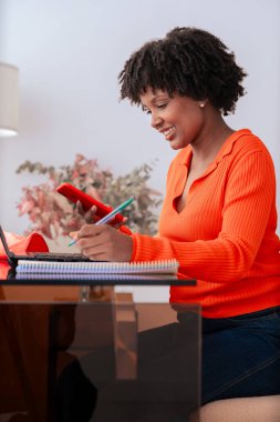 Evde eğitim gören Afro-Amerikan öğrenci, dizüstü bilgisayar üzerinde çalışırken akıllı telefon kullanıyor. Kitaplar, kırtasiye ve dekorasyonlu modern çalışma alanı. Çevrimiçi öğrenme, eğitim ve üretkenlik