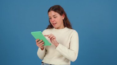 Şaşırmış bir ifadeyle, tablete yazı yazan genç bir kadın. Bir süveter giyerek, düz mavi bir arkaplan karşısında duruyor, cihazla etkileşime girerken ekrana odaklanıyor.