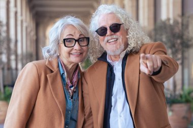 Dışarıda mutlu son sınıf çifti. Kıvırcık beyaz saçlı ve güneş gözlüklü adam kamerayı işaret ediyor, gülümsüyor, gözlüklü kadın ise yanında duruyor, hem neşeli hem de kendinden emin bir şekilde..