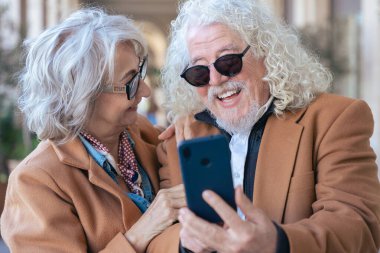 Gri saçlı ve gözlüklü yaşlı çift açık havada bir akıllı telefonu paylaşıyor. Gülüyorlar ve teknolojinin tadını çıkarıyorlar. Zarif bir şehir ortamında oturuyorlar. Kahverengi paltolarını giyip mutlu görünüyorlar..