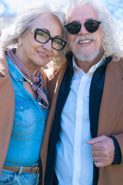 Gri saçlı ve gözlüklü yaşlı bir çift kameraya gülümsüyor. Adam güneş gözlüğü takıyor ve ikisi de zarif bir şekilde kahverengi paltolar giyip güneşli bir günün tadını çıkarıyor.. 