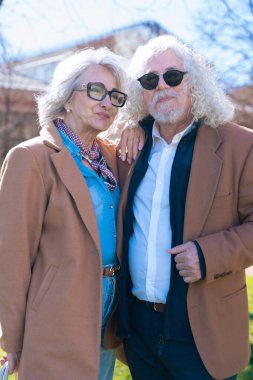 Gri saçlı ve gözlüklü yaşlı bir çift kameraya gülümsüyor. Adam güneş gözlüğü takıyor ve ikisi de zarif bir şekilde kahverengi paltolar giyip güneşli bir günün tadını çıkarıyor.. 