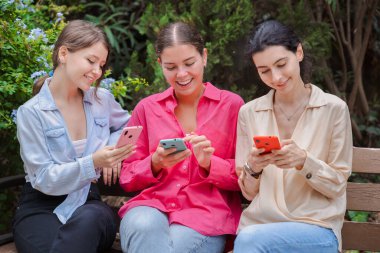 Üç beyaz kadın yeşil bir parkta ahşap bir bankta oturuyor, akıllı telefonlar kullanırken gülümsüyorlar. Mutlu ve meşgul görünüyorlar, modern cihazlar ve sosyal medya uygulamalarıyla açık havada eğleniyorlar.