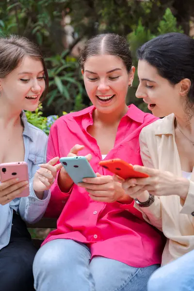 Üç beyaz kadın yeşil bir parkta ahşap bir bankta oturuyor, akıllı telefonlar kullanırken gülümsüyorlar. Mutlu ve meşgul görünüyorlar, modern cihazlar ve sosyal medya uygulamalarıyla açık havada eğleniyorlar.