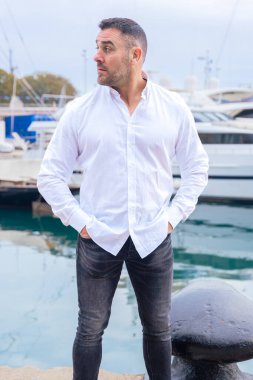 Kısa siyah saçlı ve sakallı beyaz gömlekli beyaz bir adam lüks yatların yanında duruyor. Ciddi ve kendinden emin görünüyor, yat limanında tekne ve suyla uzaktan bakıyor..