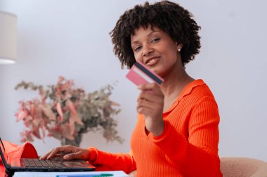 Afrikalı Amerikalı öğrenci evde dizüstü bilgisayar kullanırken kredi kartıyla çevrimiçi ödeme yapıyor. Kitaplı, kırtasiyeli ve şık dekorlu modern çalışma alanı. E-ticaret, dijital işlem ve finansman