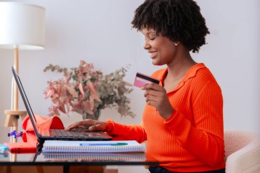 Afrikalı Amerikalı öğrenci evde dizüstü bilgisayar kullanırken kredi kartıyla çevrimiçi ödeme yapıyor. Kitaplı, kırtasiyeli ve şık dekorlu modern çalışma alanı. E-ticaret, dijital işlem ve finansman