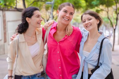 Üç neşeli genç bayandan oluşan bir grup birlikte yürüyorlar, gülümsüyorlar, gülüyorlar ve birbirlerine sarılıyorlar, rahat ve canlı bir şehir ortamında arkadaşlıklarının tadını çıkarıyorlar..