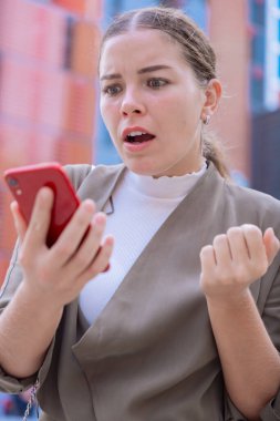 Kafkasyalı kadın şok olmuş ve şaşırmış bir ifadeyle akıllı telefonuna bakıyor. Beklenmedik haberler, sosyal medya tepkisi ve modern binaların bulunduğu kentsel bir ortamda teknoloji kavramı.