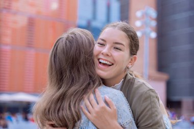  Dışarıda sarılıp gülümseyen, neşe ve dostluğu ifade eden iki mutlu beyaz kadın. Sıcak doğal ışıkla modern şehir ortamında duygusal birleşme, bağlantı ve olumlu duygular.. 