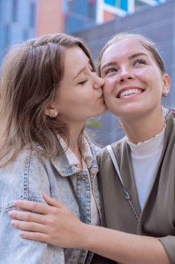 İki mutlu Kafkasyalı kadın, yanaklarından öpücükler kondurarak sevgilerini ifade ediyorlar. Arkadaşlığın, sevginin ve güvenin sembolü. Doğal ışık ve neşeli ifadelerle modern bir şehir ortamında..