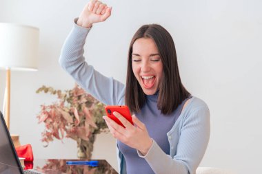 Ofis masasında oturan heyecanlı beyaz kadın, neşeyle akıllı telefona bakıyor. Modern çalışma alanında dizüstü bilgisayar, defter ve dekor. Mutluluk tepkisi, teknoloji, iletişim ve dijital etkileşim