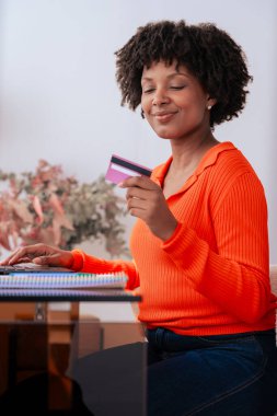 Afrikalı Amerikalı öğrenci evde dizüstü bilgisayar kullanırken kredi kartıyla çevrimiçi ödeme yapıyor. Kitaplı, kırtasiyeli ve şık dekorlu modern çalışma alanı. E-ticaret, dijital işlem ve finansman