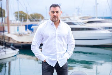 Beyaz gömlekli, kısa saçlı ve sakallı beyaz bir işadamı lüks yatların yanında duruyor. Ciddi ve kendinden emin görünüyor, yat limanında tekne ve suyla uzaktan bakıyor.
