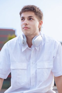 Boynuna beyaz gömlek ve kulaklık takmış genç beyaz bir adam düşünceli bir şekilde başka tarafa bakıyor. Günışığı ile aydınlık bir şehir atmosferinde modern yaşam tarzı, moda ve müzik titreşimleri..