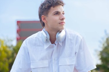 Boynuna beyaz gömlek ve kulaklık takmış genç beyaz bir adam düşünceli bir şekilde başka tarafa bakıyor. Modern yaşam tarzı, moda ve müzik atmosferi güneşli açık şehir atmosferinde.