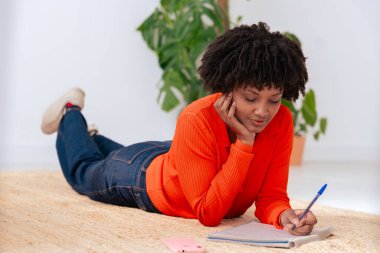 Turuncu kazaklı, rahat Afro-Amerikan kadın halılı zemine uzanıyor ve bir deftere yazı yazıyor. Arka planda yeşil bitki olan huzurlu ev ortamları, günlük ve yaratıcı kapalı alan atmosferi. 