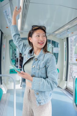 Toplu taşıma aracında dururken gülümseyen mutlu genç Çinli kadın. Kot ceket giyer ve tırabzanı tutar. Neşeli ve rahat bir tavırla yolculuktan zevk alır.. 