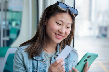 Genç Çinli kadın internet üzerinden akıllı telefonu ve kredi kartıyla toplu taşıma için ödeme yapıyor. Pencerenin kenarında otururken gülümsüyor, iş seyahati sırasında düzgün bir dijital işlemin tadını çıkarıyor..