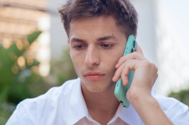 Kafkasyalı genç adam akıllı telefonuyla ciddi bir ifadeyle konuşuyor. Açık havada beyaz bir gömlek giyiyor, odaklanmış ve düşünceli bir şekilde doğal bir şehir ortamında sohbet ediyor.. 