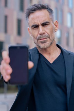 Siyah kıyafetli, kendine güvenen beyaz adam kameraya doğru ciddi bir yüzle akıllı telefon tutuyor. Modern mimariyle kentsel bir çevrede iş, teknoloji ve iletişim kavramı. 
