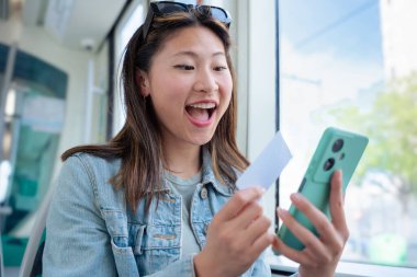 Genç Çinli kadın internet üzerinden akıllı telefonu ve kredi kartıyla toplu taşıma için ödeme yapıyor. Pencerenin kenarında otururken gülümsüyor, iş seyahati sırasında düzgün bir dijital işlemin tadını çıkarıyor..