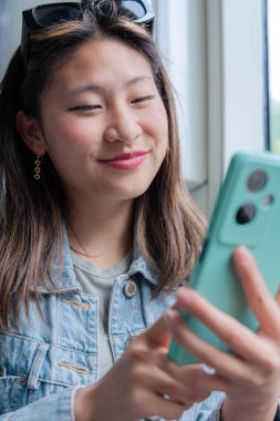 Kot ceketli Çinli kadın tramvayda oturuyor, gülümsüyor ve telefonunda bir şeyler okuyor. Şehir arkaplanı, günlük tarz, toplu taşıma, pozitif ya da mutlu ifadeler. Dikey