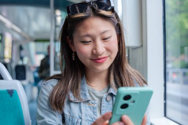 Kot ceketli Çinli kadın tramvayda oturuyor, gülümsüyor ve telefonunda bir şeyler okuyor. Şehir arkaplanı, günlük tarz, toplu taşıma, pozitif ya da mutlu ifadeler