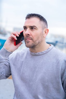 Kafkasyalı bir adam suyun yanında duruyor, akıllı bir telefonla ciddi ciddi konuşuyor. Kıyafeti ve yoğun ifadesi önemli bir telefon konuşması sırasında yoğunlaştığını gösteriyor. Dikey