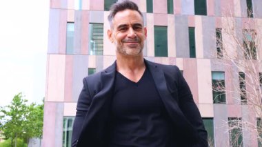 Kafkas işadamı elleri cepte, modern bir iş bölgesinde kameraya bakıyor. Gülümseyen ifadesi ve kendine güveni başarıyı, liderliği ve kararlılığı yansıtıyor..