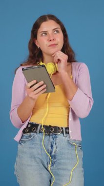 Kafkas bir kadının dikey portresi, düşünceli bir yüz ifadesi var, elinde tablet ve sarı kulaklık takıyor. Gelişigüzel stil, mavi arkaplan ve dijital odak modern bağlantıyı akla getiriyor. Dikey