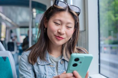 Kot ceketli Çinli kadın tramvayda oturuyor, gülümsüyor ve telefonunda bir şeyler okuyor. Şehir arkaplanı, günlük tarz, toplu taşıma, pozitif ya da mutlu ifadeler